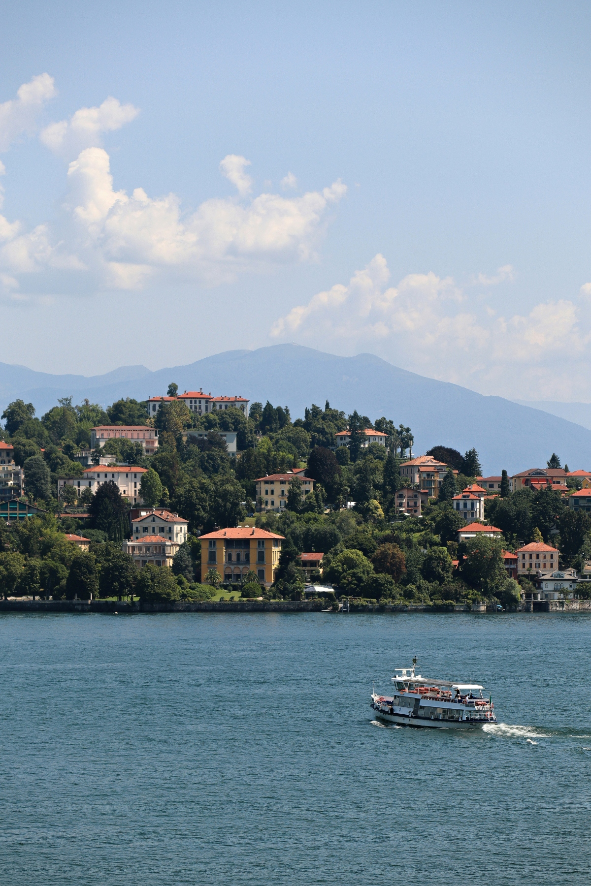 A passenger ferry crosses blue lake waters in the foreground, with a densely built hillside town rising behind it featuring multi-story buildings with terracotta roofs nestled among green trees and vegetation. Hazy blue mountains form the backdrop beneath a partly cloudy sky.
