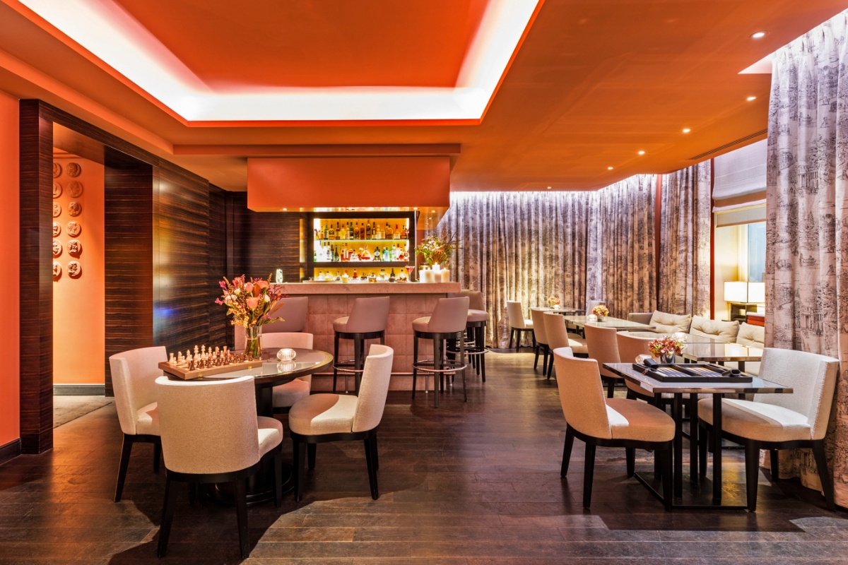 Hotel bar and lounge featuring a bold orange ceiling with recessed lighting and a central bar area with illuminated shelving displaying bottles and glassware, set against dark horizontal wood paneling. The space includes dark wood flooring and multiple seating areas with cream-colored upholstered chairs arranged around low tables displaying chess sets and board games, with floral arrangements. Floor-to-ceiling patterned curtains line the right side of the room, separating the lounge from adjacent spaces with additional seating visible in the background.