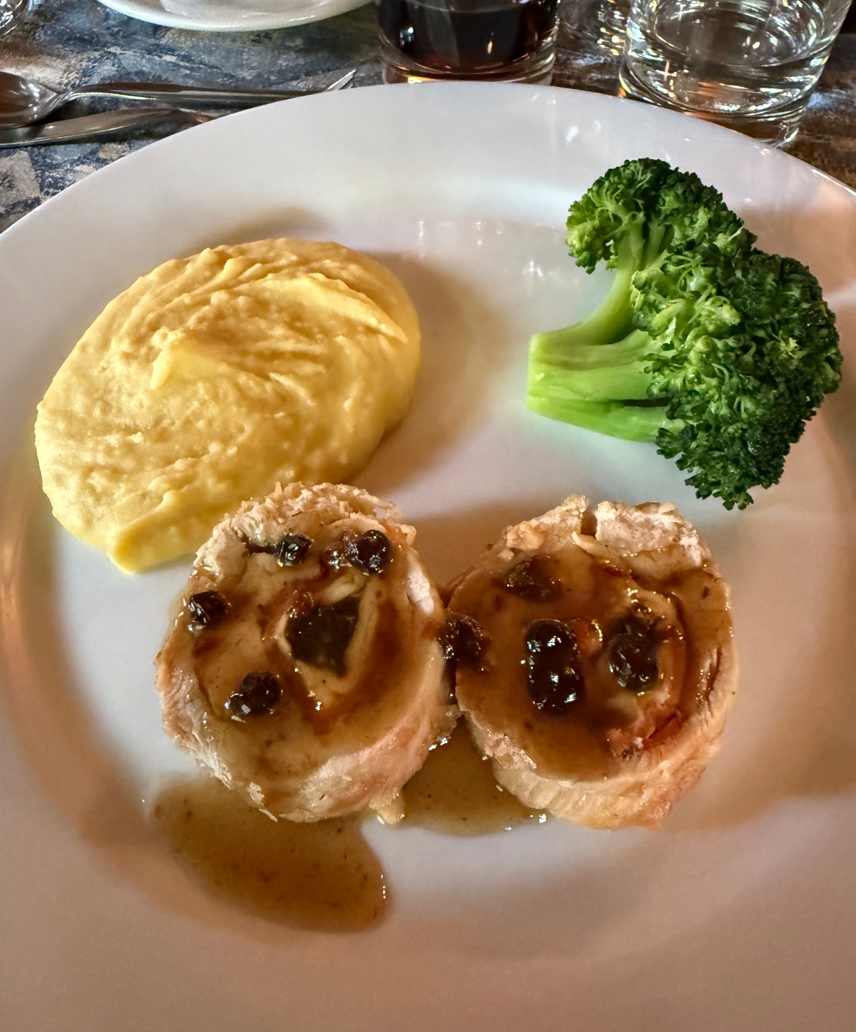 A white plate presents two sliced portions of stuffed meat topped with a dark caramelized sauce, accompanied by a smooth mound of golden mashed potatoes and bright green steamed broccoli. The plated meal sits on a dark stone surface with drinking glasses and silverware visible in the background.