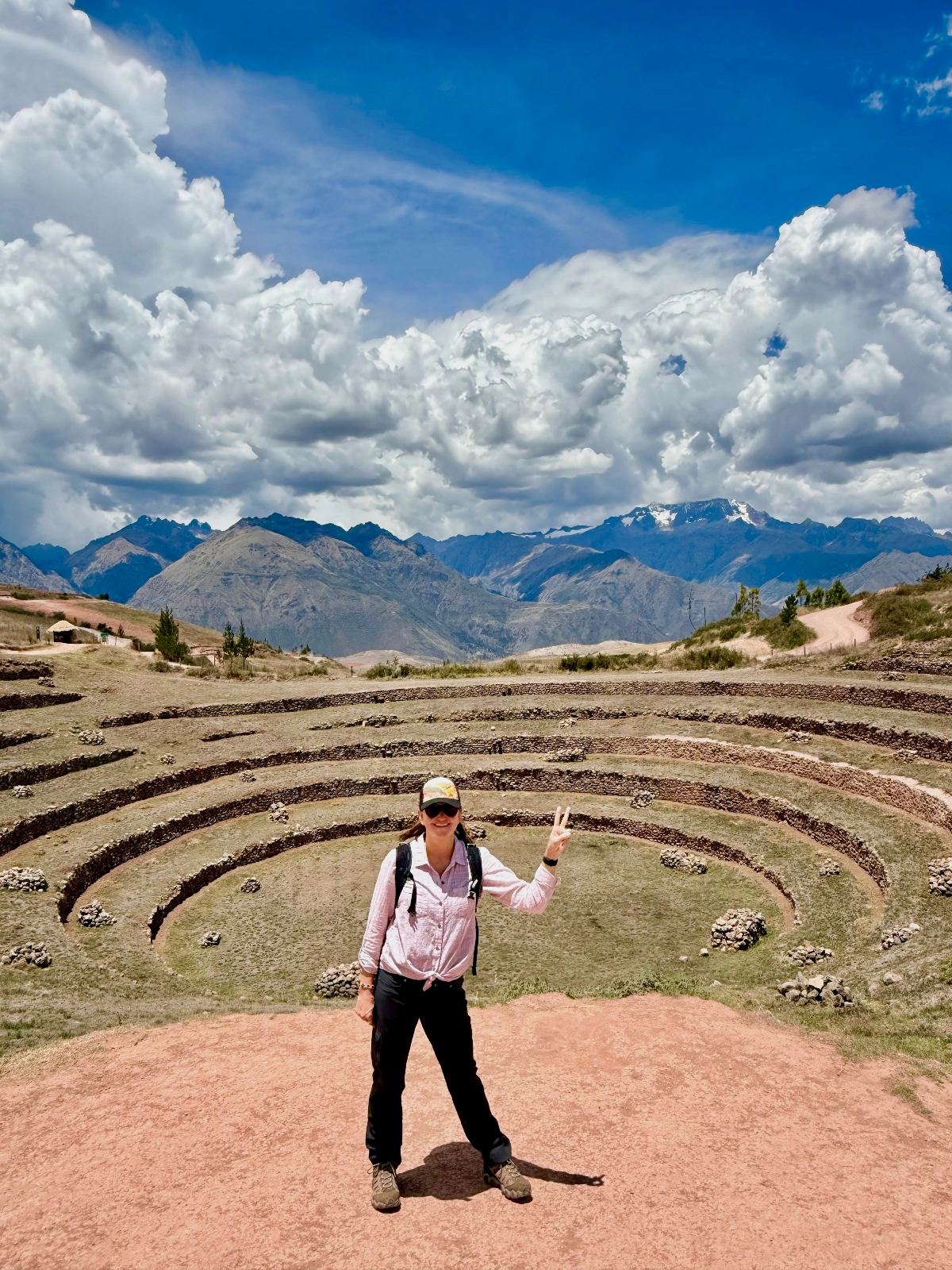 A woman in hiking attire wearing a baseball cap, sunglasses, and backpack makes a peace sign while standing on a dirt path at the edge of circular concentric terraced ruins carved into the earth. Behind her, the ancient agricultural terraces descend in rings against a backdrop of snow-capped Andean mountains and dramatic white clouds in a bright blue sky.
