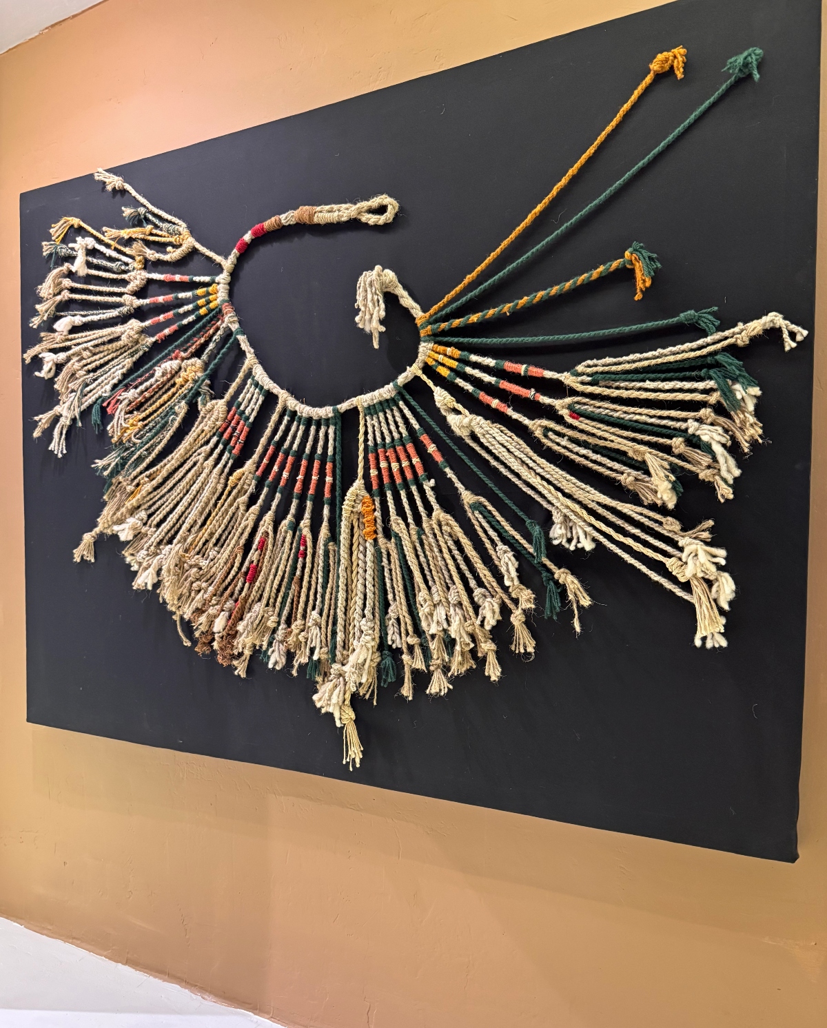 A museum display shows an ancient knotted cord recording device mounted on a black board, consisting of a central rope from which dozens of colored strings hang with knots tied at various intervals along their length. The strings feature tasseled ends and display a palette of natural tan, coral orange, dark green, black, and yellow fibers in geometric patterns, with additional twisted cords in yellow and green extending from the top.