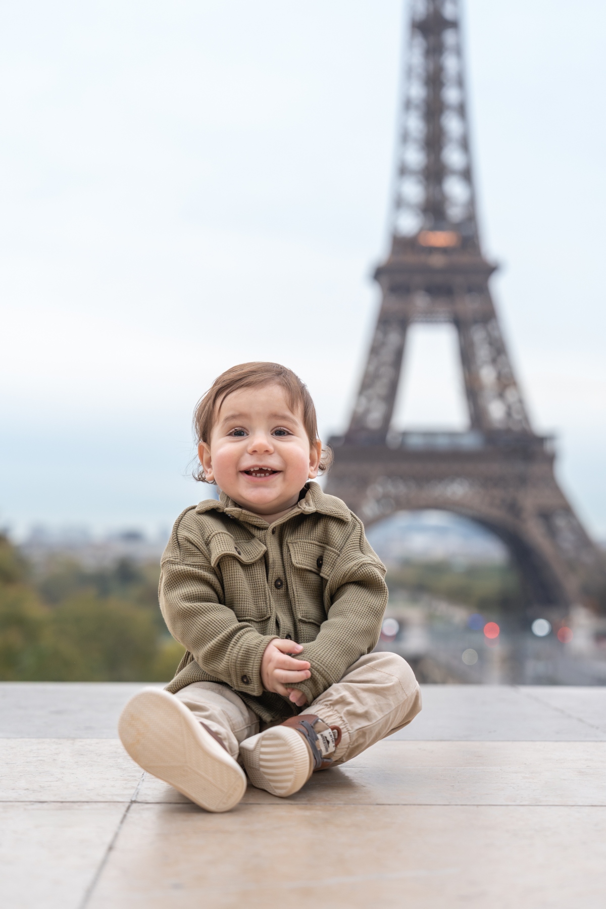 A toddler sits cross-legged on a stone plaza surface wearing an olive-green textured jacket and beige pants with white sneakers. The Eiffel Tower stands out of focus in the background against an overcast sky, with the viewing area and distant cityscape visible behind the child.