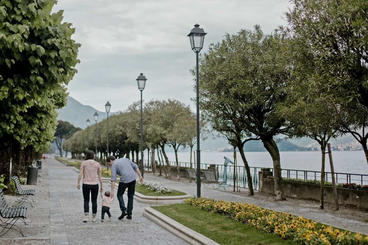 Two adults walk hand-in-hand with a toddler along a tree-lined lakeside promenade under an overcast sky. The paved walkway features ornamental street lamps, benches, flower beds with yellow blooms, and views of a large lake with mountains visible in the distance.