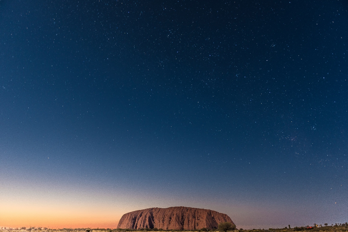 A large sandstone monolith with vertical striations rises from flat desert terrain under a twilight sky that transitions from warm orange and peach tones near the horizon to deep blue filled with visible stars. Sparse vegetation dots the surrounding landscape, and the expansive sky occupies approximately three-quarters of the vertical frame.
