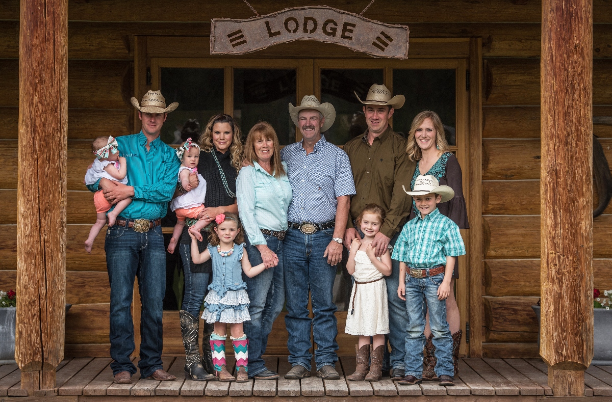 A multi-generational group of ten people poses on the wooden porch of a log cabin with a metal "LODGE" sign hanging above the entrance. The group wears Western attire including cowboy hats, pearl snap shirts, jeans, and boots, arranged in two rows with adults in back and children in front.