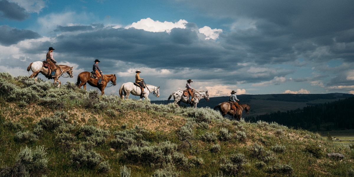 Five horseback riders in western attire traverse a sagebrush-covered hillside against a backdrop of dramatic storm clouds. The riders, mounted on horses of varying colors including white and brown, are silhouetted against distant mountain ranges and forested valleys.