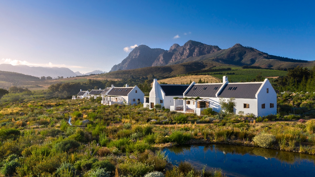 A cluster of traditional white-washed buildings with dark pitched roofs sits amid cultivated gardens and natural vegetation, with a reflecting pond in the foreground. Dramatic rocky mountains rise behind the farm complex under a clear blue sky, with vineyard rows and agricultural land visible across the surrounding hillsides.
