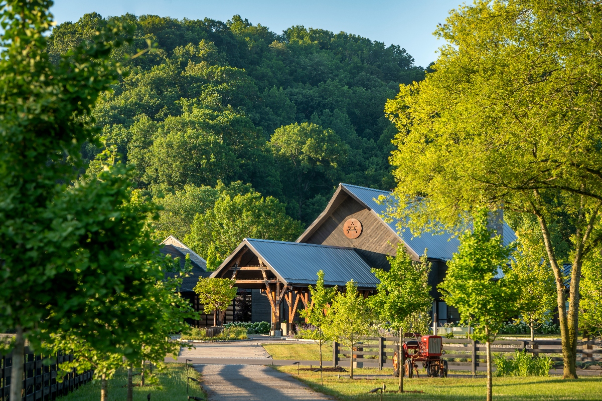 A rustic wooden barn with a metal roof and circular emblem sits at the end of a tree-lined driveway, framed by dense forest and rolling hills in the background. The pastoral setting features wooden post fencing in the foreground, a vintage red farm wagon, and mature trees casting late afternoon light across the property.