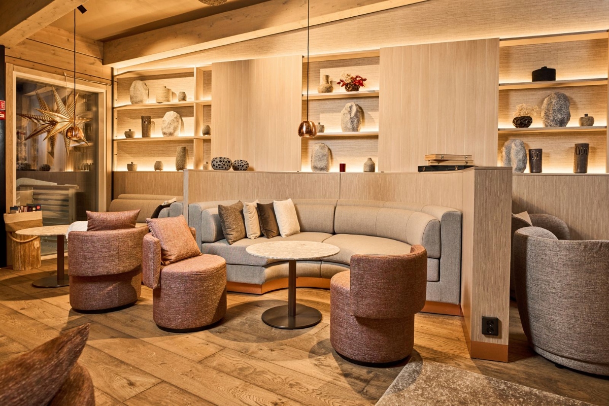 A modern hotel lobby features light wood flooring and a seating area with a curved gray sofa, round upholstered ottomans in mauve-pink tones, and a small circular coffee table. The back wall consists of floor-to-ceiling light wood shelving units with integrated LED lighting, displaying various decorative objects and sculptures, with a reception desk visible on the right side.
