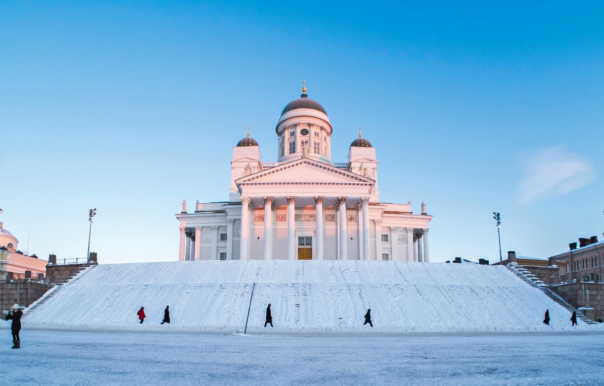 A white neoclassical building with a central green dome and two smaller flanking domes sits atop a large snow-covered staircase, its columned portico illuminated against a clear blue sky. Several people walk across the snow-covered plaza in front of the building, appearing as small dark figures against the white snow.