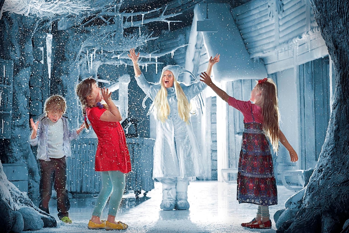 Three children and a character dressed in white winter costume with long blonde hair and pointed ears stand in an indoor ice cave attraction decorated with icicles and snow-covered tree trunks. Artificial snow falls throughout the blue-lit space as the children reach up toward the falling snow with raised hands.