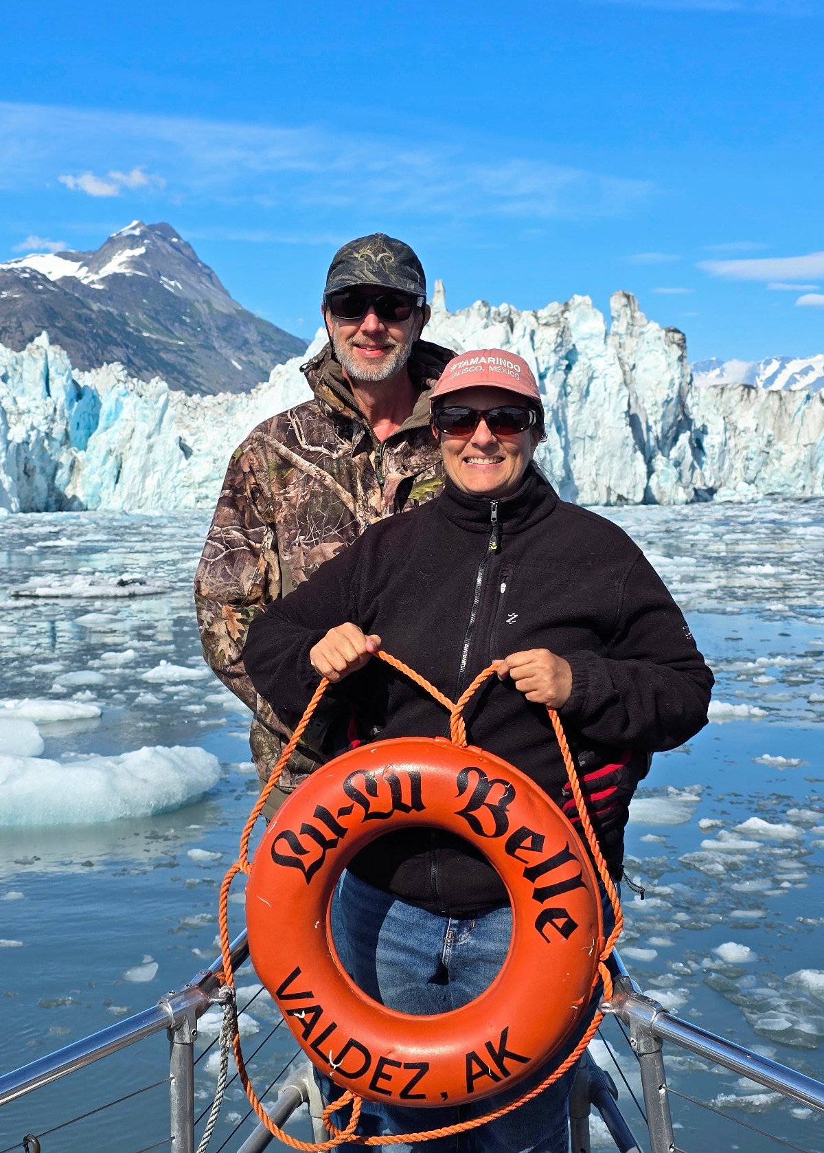 Two people stand on the stern deck of a boat called the Lu-Lu Belle out of Valdez, Alaska, posing with an orange life preserver ring while floating among ice chunks in glacier-dotted waters. A massive tidewater glacier face and snow-capped mountains rise dramatically under a bright blue sky in the background.