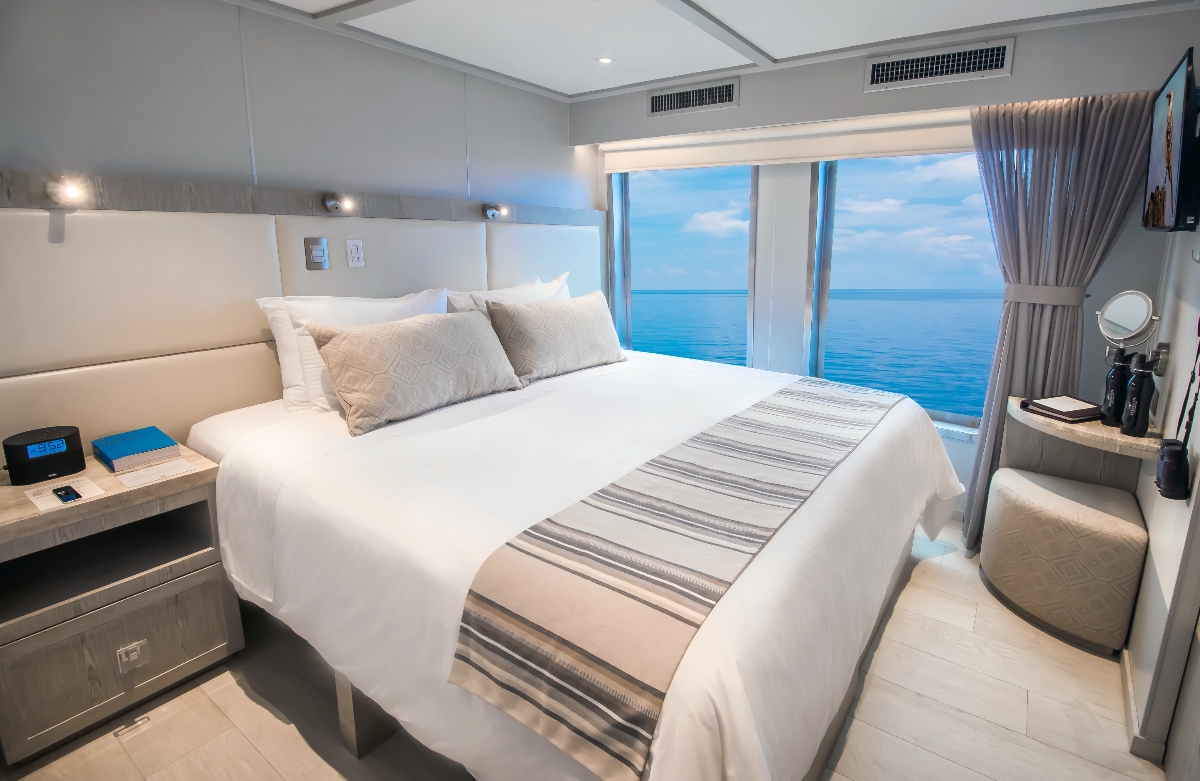 A modern ship cabin features a king bed dressed in crisp white linens with a woven stripe runner, flanked by a wood nightstand with a clock and reading lamp. Floor-to-ceiling windows along the back wall frame a panoramic view of the open ocean under a blue sky, with a small seating area and vanity visible to the right.