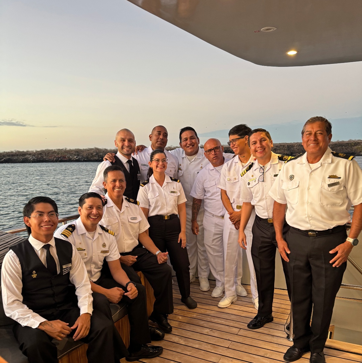A group of approximately eleven smiling ship crew members in white uniforms and formal service attire pose together on a teak deck at dusk. A rocky Galápagos coastline and calm water are visible in the soft evening light behind them.