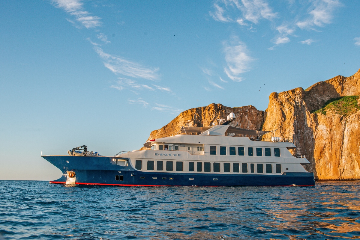 The Ecoventura Evolve, a sleek blue and white luxury expedition vessel, sits anchored on calm blue water in warm golden light. Towering amber-colored volcanic cliffs rise dramatically behind the ship against a clear blue sky with wispy clouds.