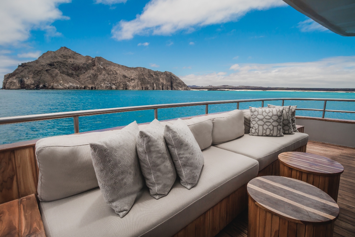 A cushioned outdoor sofa with patterned throw pillows sits on a teak deck alongside two round wood side tables, overlooking a chrome railing and vivid turquoise water. A rugged volcanic island rises from the sea in the middle distance beneath a bright blue sky with scattered clouds.