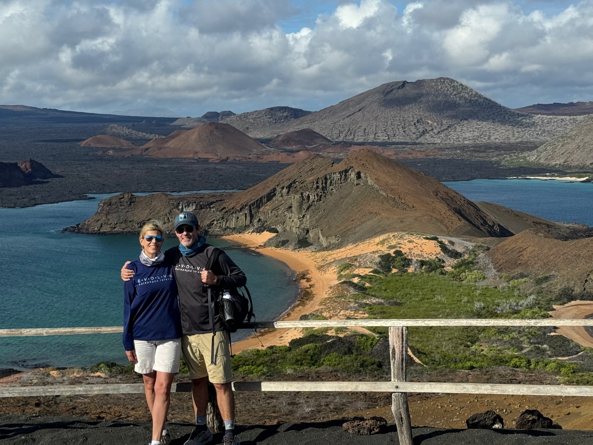 Two adults wearing Galápagos-branded athletic wear pose together at a wooden railing overlook, with a sweeping panorama of volcanic cinder cones, a golden sand beach, and turquoise bays stretching behind them. Multiple rust-colored and gray volcanic peaks rise across the landscape under a partly cloudy sky, showcasing the dramatic, otherworldly terrain of the Galápagos Islands.