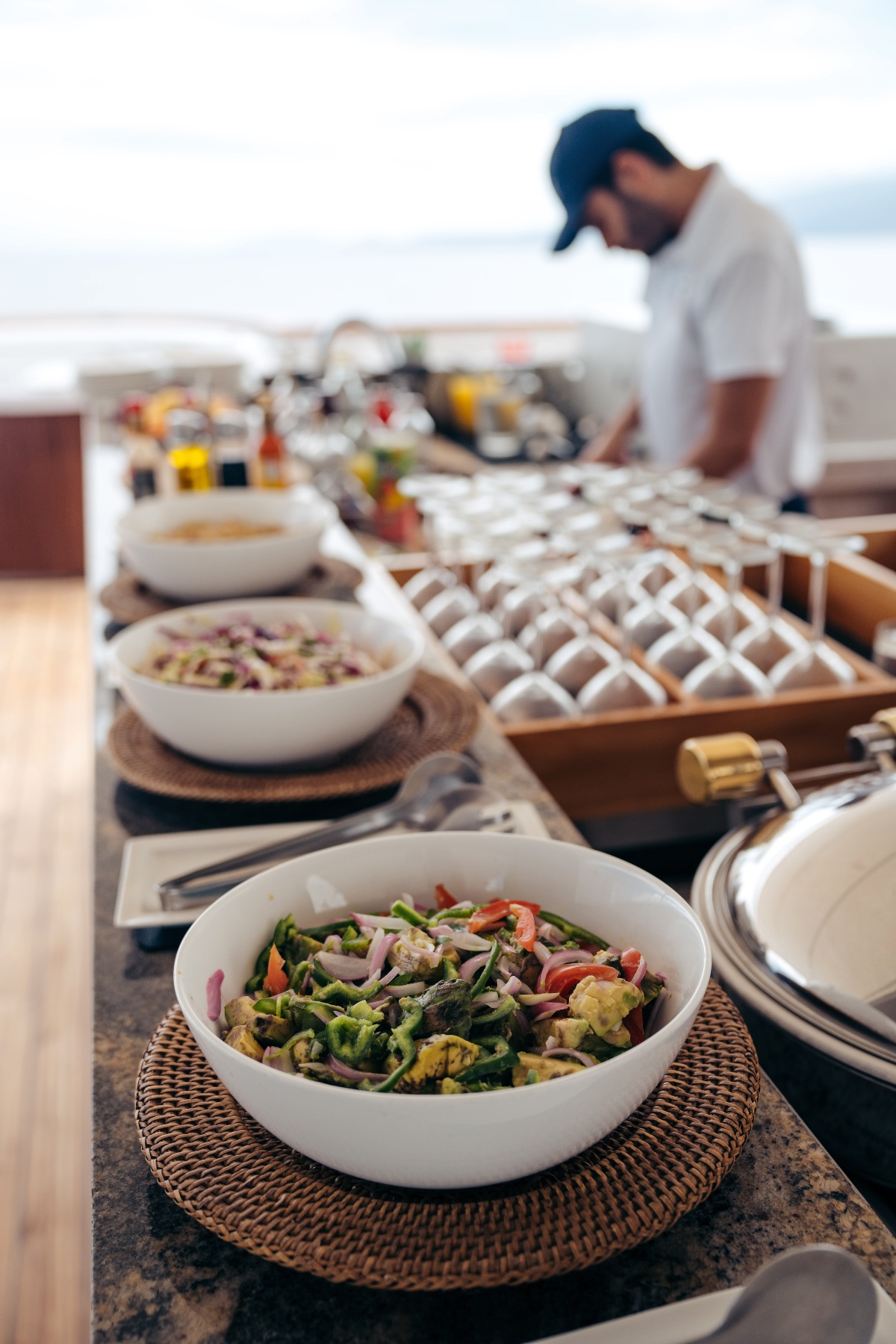 A buffet spread on an outdoor deck features white bowls of fresh vegetable salads on woven rattan chargers, with a rack of stemware and chafing dishes extending along the table. A crew member in a white shirt and navy cap tends to the buffet in the background, with the open ocean visible beyond.