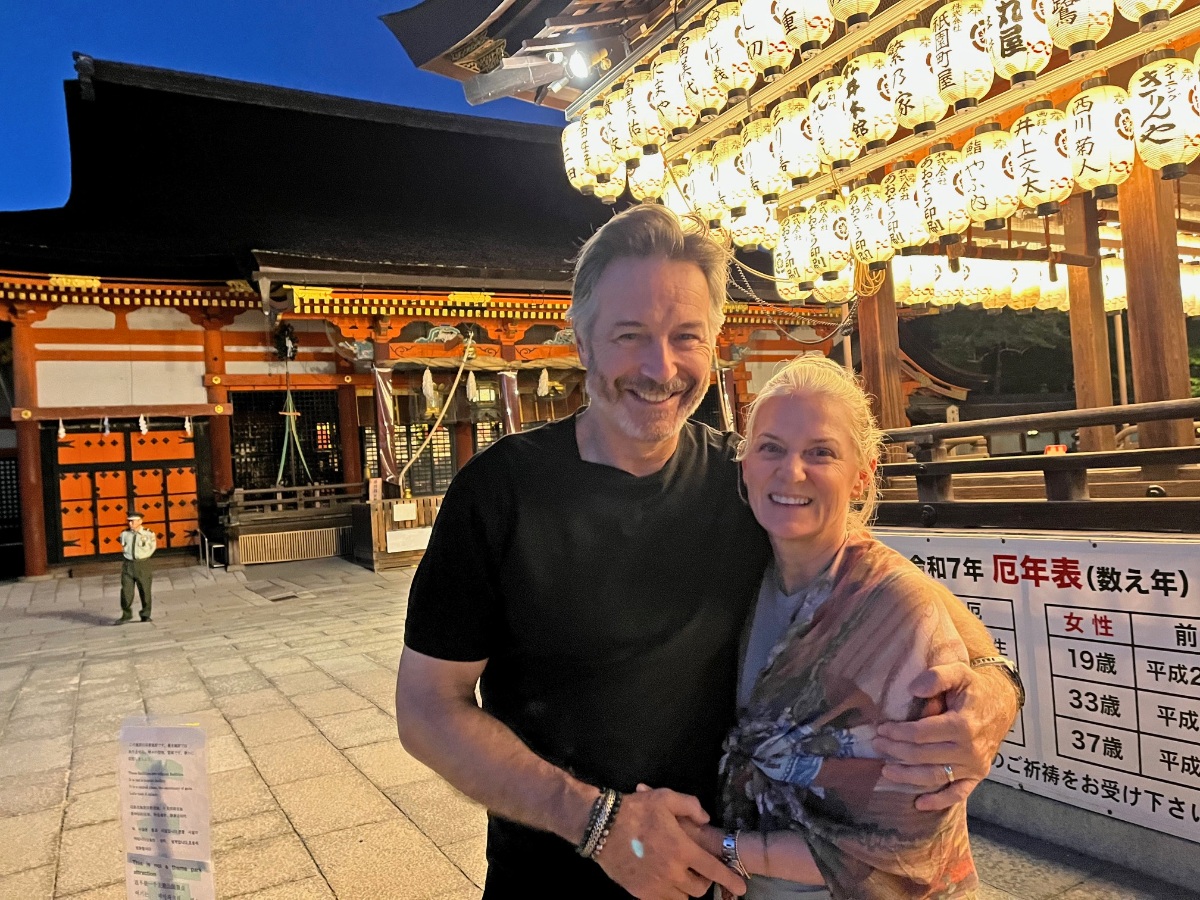 Two smiling adults pose together in front of a traditional Japanese shrine at dusk, surrounded by rows of glowing white paper lanterns bearing Japanese kanji inscriptions. The ornate shrine building features vibrant orange and red architectural details, while a Japanese signboard listing unlucky years (yakudoshi) is visible to the right.