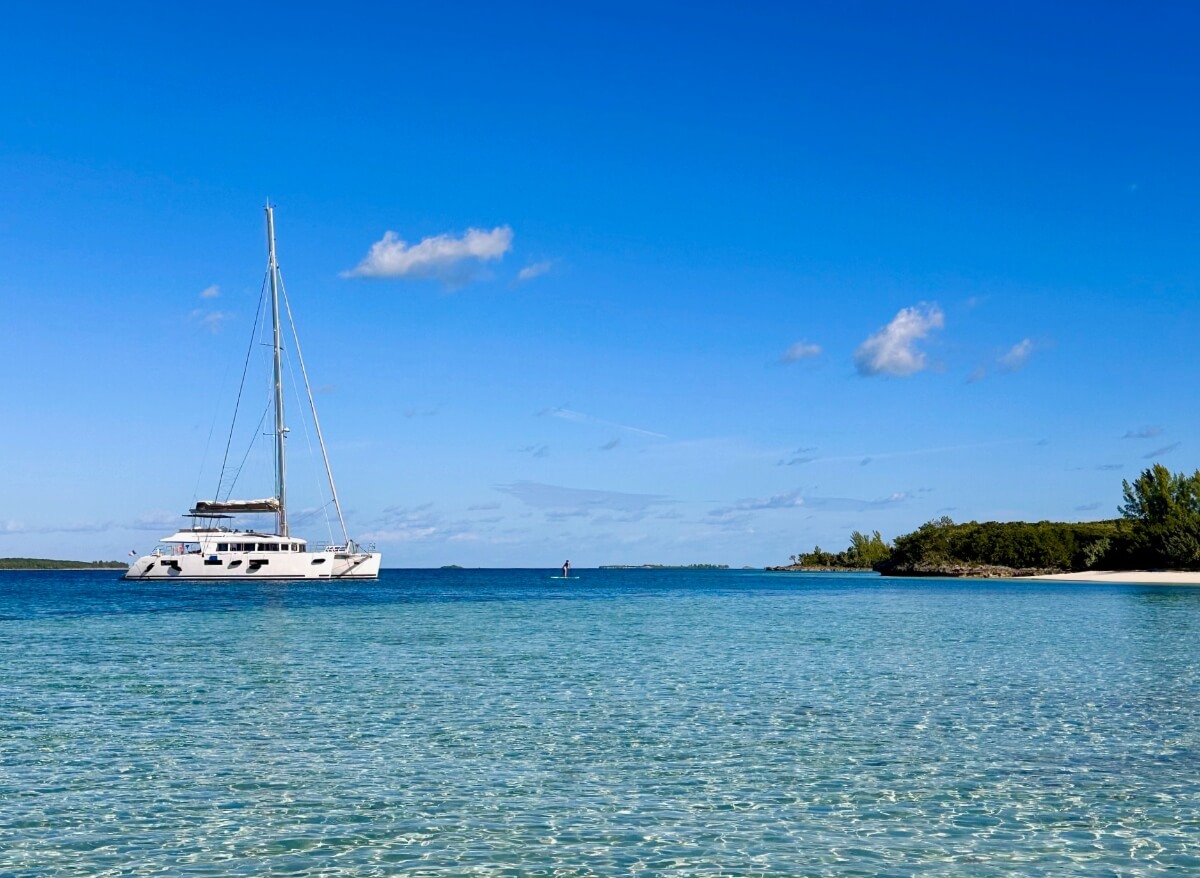 A white catamaran with a tall mast is anchored in calm turquoise water beneath a bright blue sky with scattered white clouds. A tree-lined shore with a sandy beach is visible in the distance to the right.