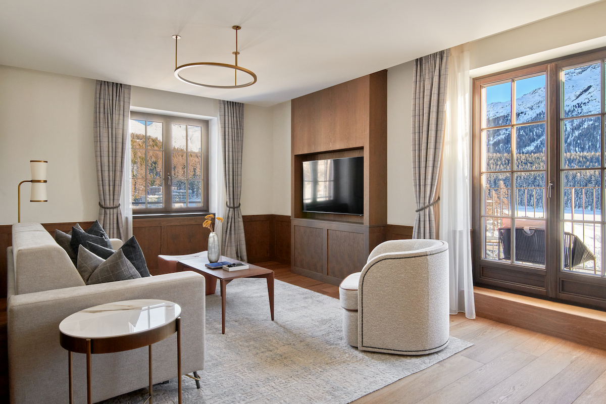 A modern living room features light wood flooring, wood-paneled walls with a mounted television, and neutral-toned furniture including a sofa, barrel chair, and side tables beneath a circular brass pendant light. Two windows with gray curtains frame views of snow-covered mountains, with one window opening to a balcony through French doors.