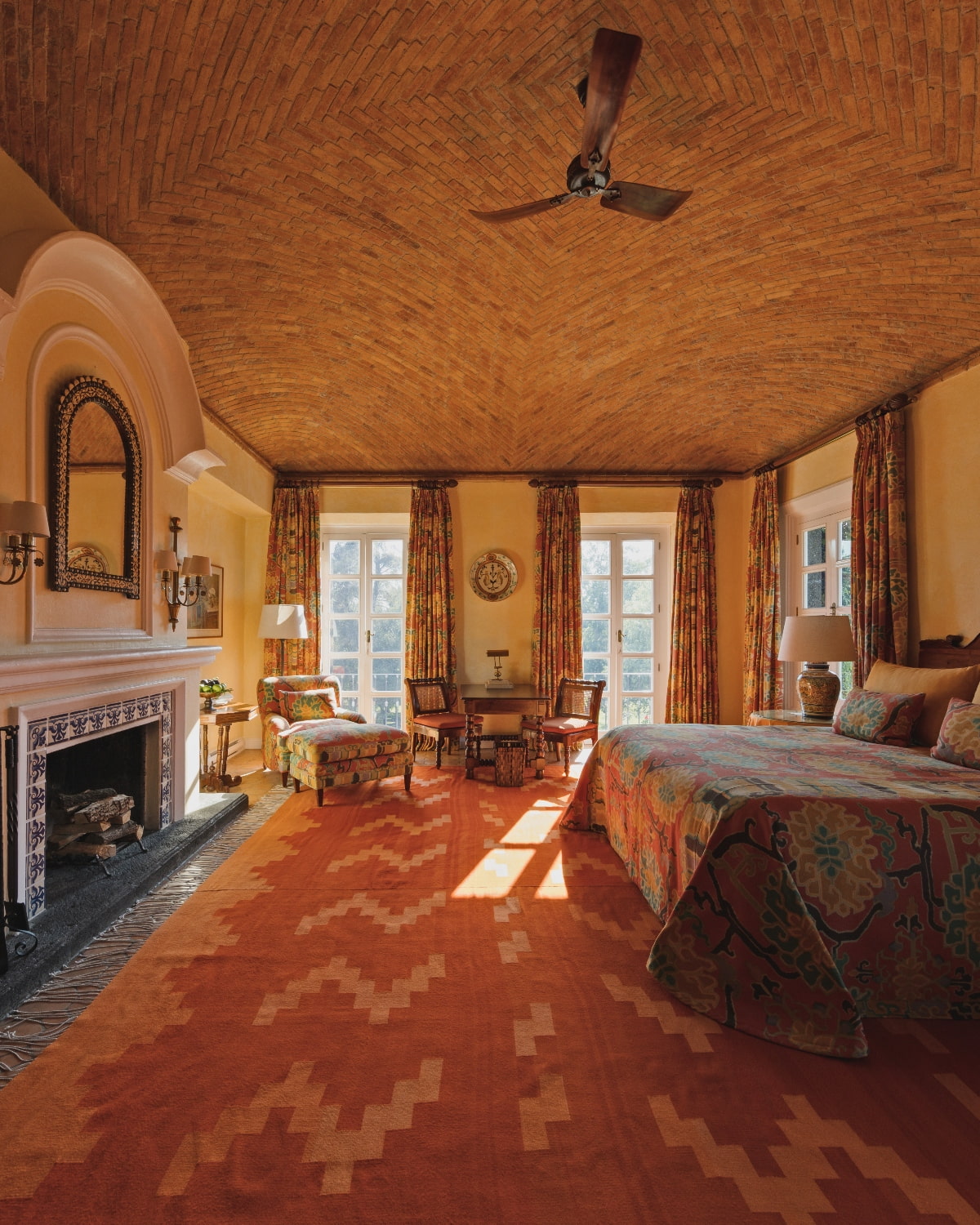 A spacious bedroom features a dramatic barrel-vaulted ceiling covered in terracotta brick laid in a herringbone pattern, with a wooden ceiling fan at its center. The room includes a tiled fireplace with an arched mirror, French doors with patterned draperies, a bed with colorful bedding, upholstered seating, and geometric-patterned terracotta tile flooring.