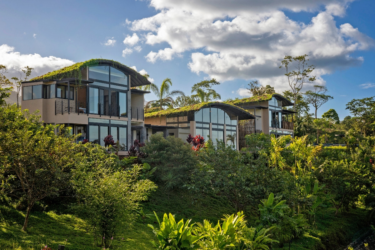 A modern multi-level building features curved barrel-vaulted roofs covered with green vegetation and large floor-to-ceiling glass windows. The structure is surrounded by lush tropical landscaping including palm trees, tropical plants, and colorful foliage beneath a partly cloudy blue sky.