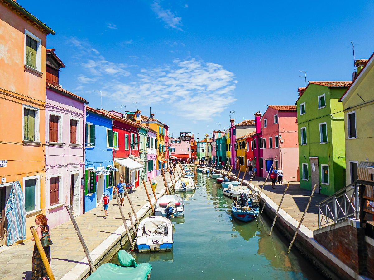 A narrow canal on the island of Burano is flanked on both sides by densely packed houses painted in vivid shades of pink, purple, blue, red, orange, and green, with small motorboats moored along the canal edges. Pedestrians walk along the sunny waterfront walkways beneath a bright blue sky, with wooden mooring poles lining both banks.