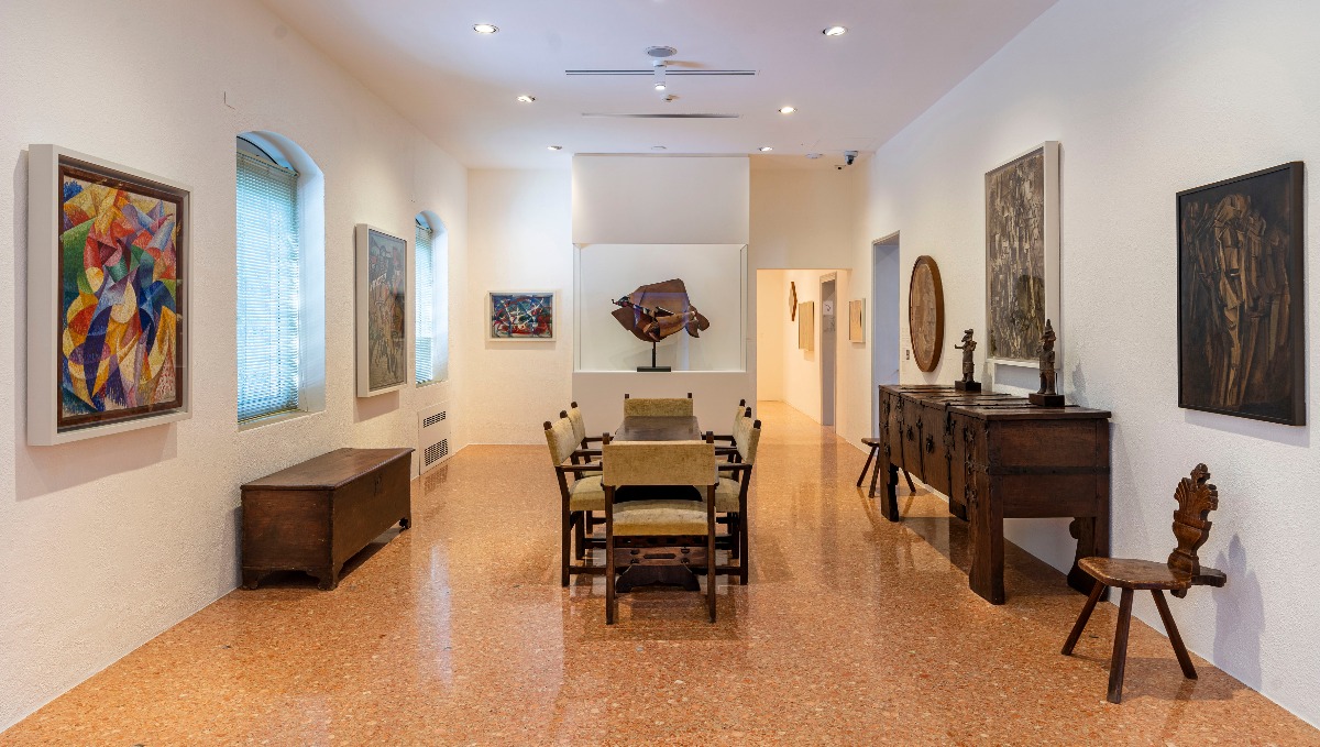 A bright gallery room at the Peggy Guggenheim Collection in Venice features white walls hung with modernist and Cubist paintings, a polished terrazzo floor, and a central dark wood dining table surrounded by upholstered chairs. Antique wooden furniture is arranged throughout the space, and a bronze abstract sculpture is displayed on a pedestal at the far end of the room.