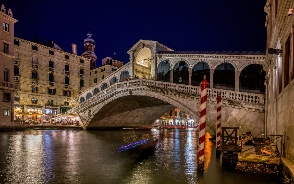 The illuminated Rialto Bridge arches over the Grand Canal at night, its white stone balustrades and colonnaded porticoes reflected in the dark water below, with a boat's light trails streaking beneath the span. Lit restaurants and historic buildings line the canal banks on both sides, and a red-and-white striped mooring pole stands in the foreground alongside a small waterside terrace.