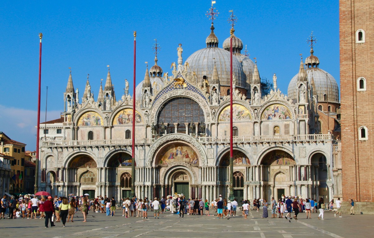 The ornate Byzantine facade of St. Mark's Basilica in Venice dominates the frame, featuring five arched portals, elaborate golden mosaics, marble sculptures, and multiple domes rising against a vivid blue sky. A large crowd of visitors fills the sun-drenched piazza in front of the cathedral, with the base of the Campanile visible at the right edge of the image.