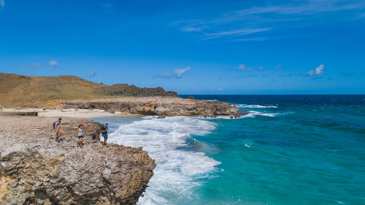 Three hikers make their way along a rugged limestone coastline where turquoise waves break against the rocks below. A sandy cove, arid hillside, and deep blue sky stretch into the distance behind them.