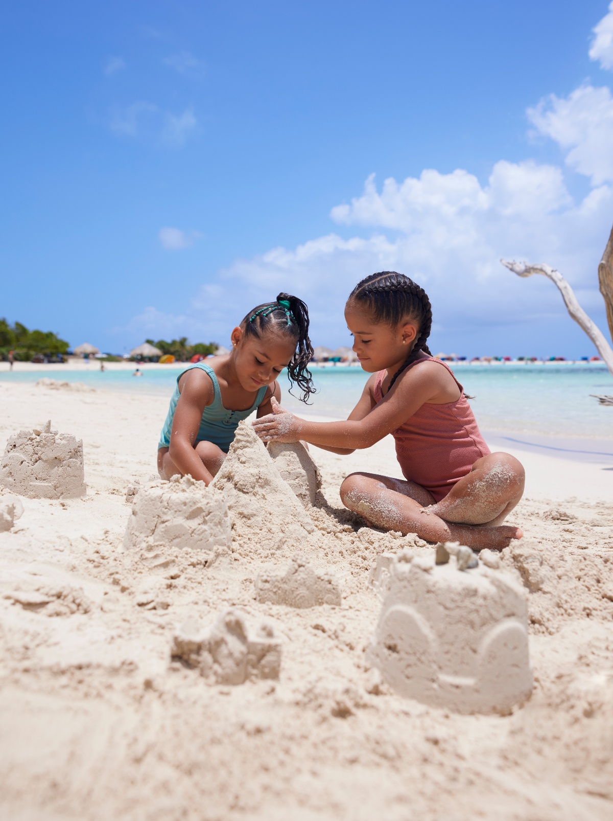 Two young girls in swimsuits crouch together on a white sand beach, building sandcastles with several small mounds of sand around them. Turquoise water, thatched beach umbrellas, and a bright blue sky are visible in the background.