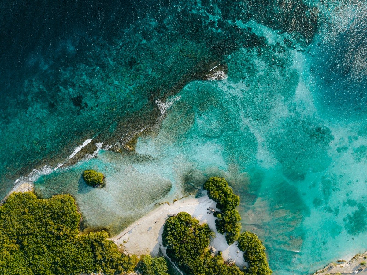 An aerial view shows a narrow sandy shoreline bordered by dense green mangroves, where shallow turquoise water grades into deeper teal and dark blue beyond a coral reef. Small waves break along the reef's edge, revealing the contrast between the protected lagoon and the open sea.