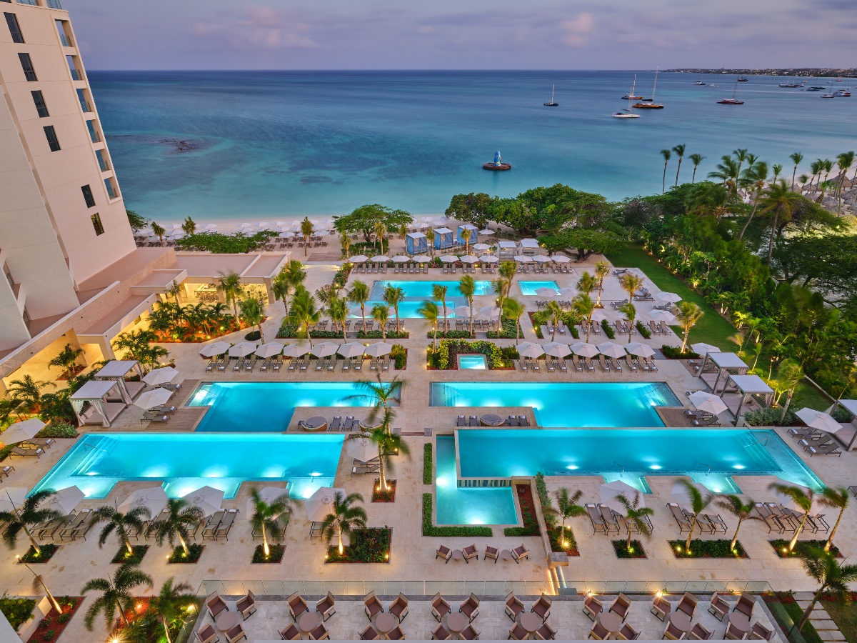 Aerial dusk view of a luxury beachfront resort featuring multiple illuminated rectangular pools surrounded by lounge chairs, palm trees, and white umbrellas on a stone terrace. The resort grounds extend to a white sand beach and calm turquoise Caribbean waters dotted with sailboats in the background.