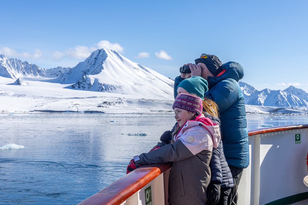 Two young girls and an adult, dressed in winter layers and hats, stand at the wooden railing of a cruise ship deck looking out over an ice-dotted Arctic waterway. The adult holds binoculars to their eyes while the girls lean against the railing, with dramatically snow-covered mountains rising steeply from the shoreline under a clear blue sky.
