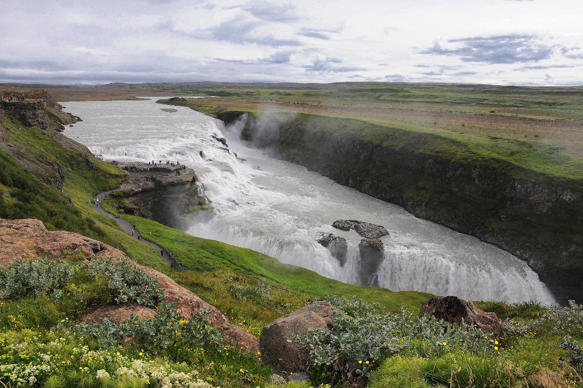A powerful two-tiered waterfall cascades into a deep rocky canyon, sending up a thick mist as the churning white water disappears into the gorge below. A winding footpath along the canyon's green mossy edge leads to a viewing platform where small figures of visitors can be seen, giving a sense of the waterfall's immense scale. Rocky outcroppings and low wildflowers fill the foreground, while a wide flat plain stretches to the horizon under an overcast sky.