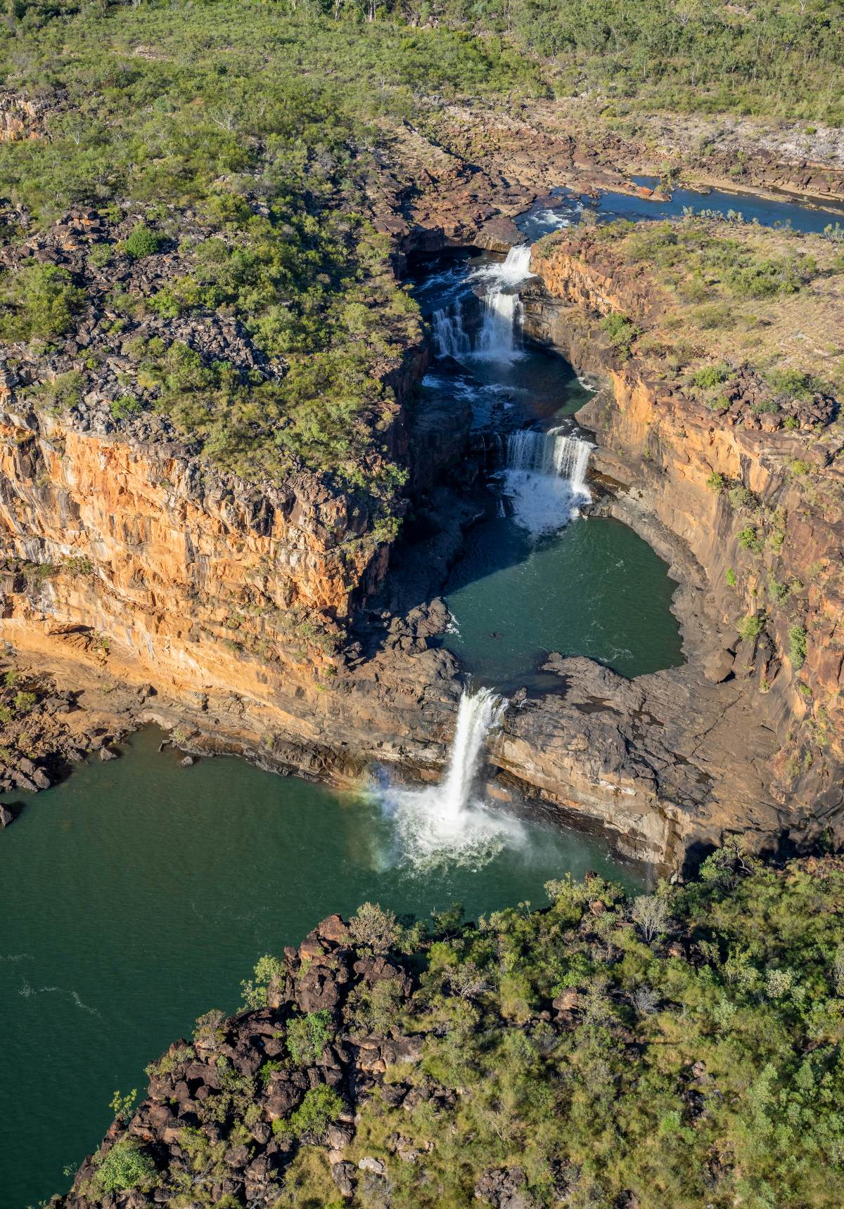 An aerial view reveals a multi-tiered waterfall system descending through a series of layered sandstone gorges, with each cascade dropping into a green pool before flowing to the next level below. The stratified rock faces display vivid bands of orange and ochre, contrasting with the deep teal of the plunge pools and the dense green bushland covering the surrounding plateau. A small rainbow is visible in the mist at the base of the final and tallest drop.