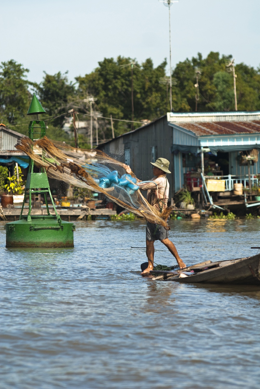 A man standing barefoot on the bow of a narrow wooden boat casts a large fishing net over the water, the net caught mid-throw in a wide arc. A green channel marker buoy sits in the river beside him, with stilt houses and corrugated metal-roofed structures lining the riverbank in the background.