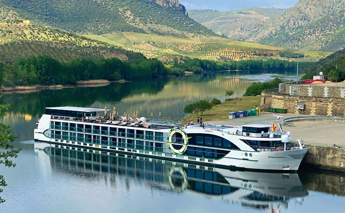 The Tauck river cruise ship Andorinha is docked alongside a stone quay on the calm Douro River, its white multi-deck hull reflected in the still water below. Terraced vineyards and olive groves cover the rolling hills in the background, with green tree-lined banks framing the river on both sides.