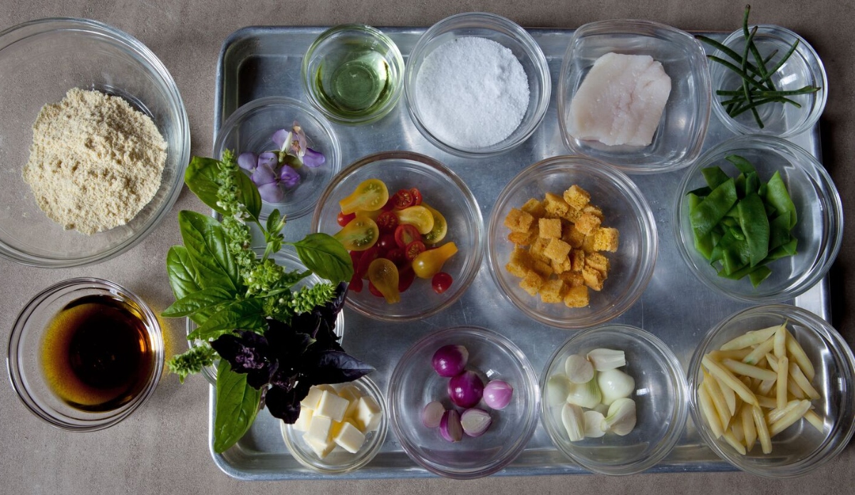 A metal baking tray holds approximately a dozen small glass bowls arranged in a grid, each containing a separate mise en place ingredient including cherry tomatoes, fresh basil, cubed croutons, coarse salt, raw fish, snap peas, pearl onions, julienned vegetables, butter, herbs, edible flowers, and a dark liquid sauce. The overhead shot is taken from directly above on a neutral gray surface.