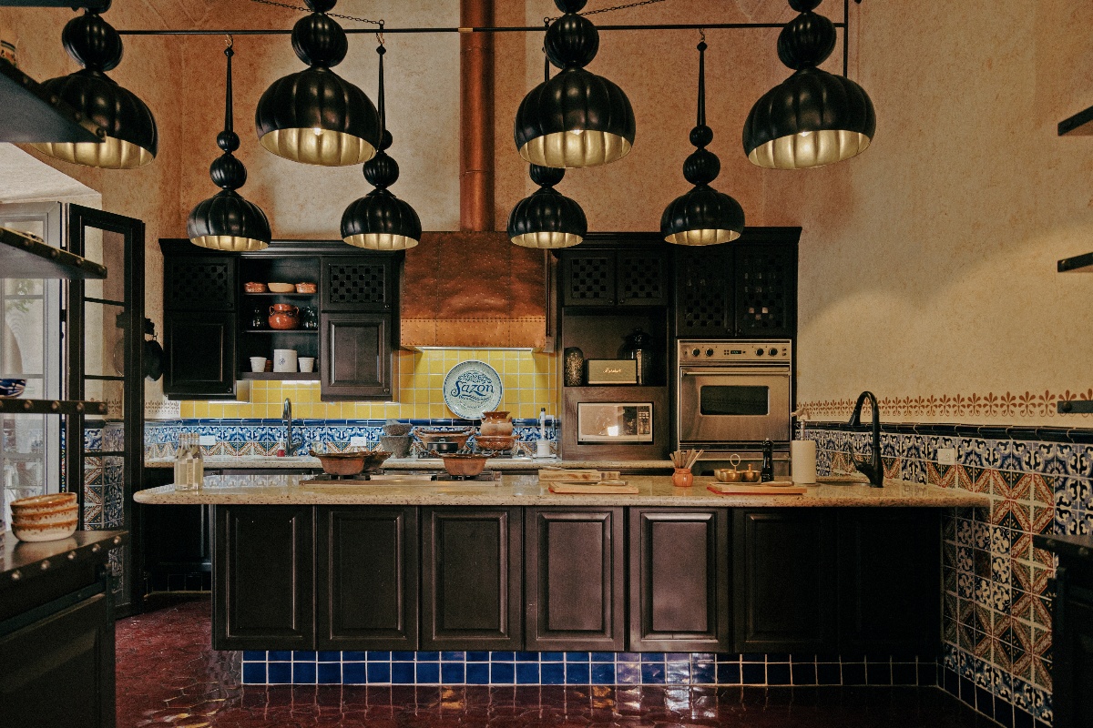 A Mexican-style kitchen features dark wood cabinetry, a stone-topped island, and a large copper range hood flanked by yellow and blue Talavera tile backsplash. Eight matte black pendant lights with gold interiors hang in two rows above the island, and patterned ceramic tiles line the walls and base of the cabinetry.