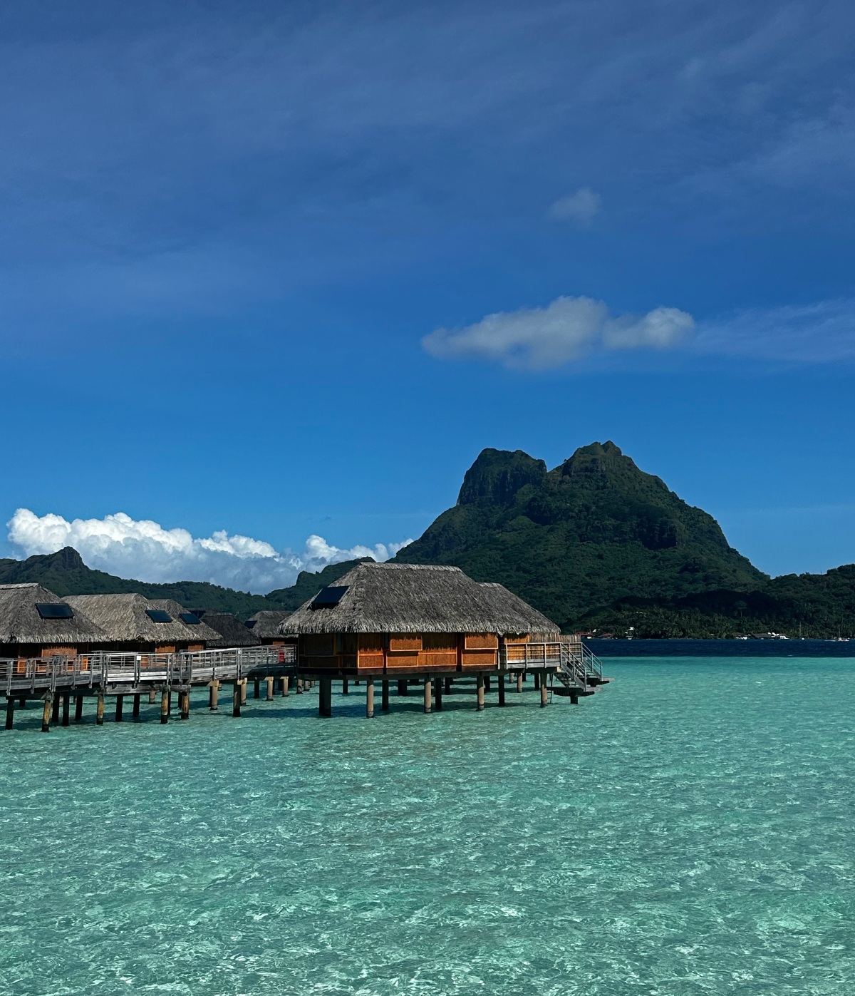 A row of overwater bungalows with thatched roofs and warm wood siding stands on wooden stilts above a clear, shallow turquoise lagoon, connected by a wooden walkway with railing. The iconic twin peaks of Mount Otemanu rise sharply against a deep blue sky in the background, their densely forested slopes dominating the horizon. The still, glassy water in the foreground reflects the bright midday light, with the lagoon's sandy bottom visible beneath the surface.