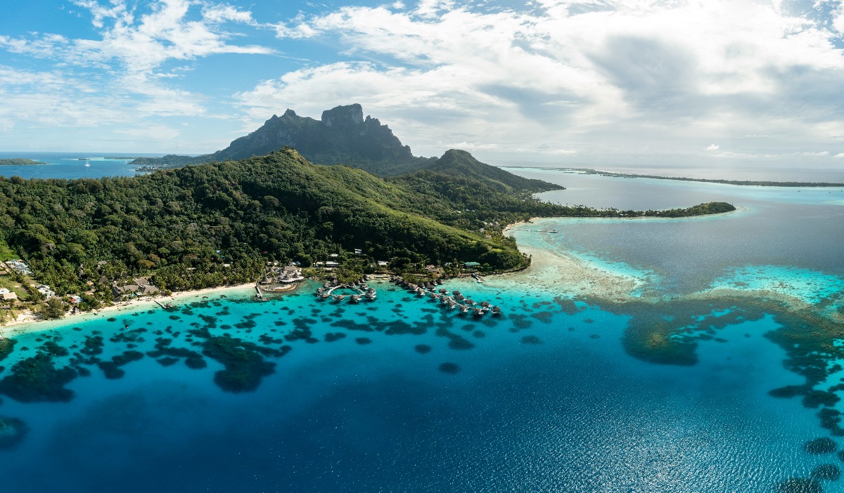 An aerial view of Bora Bora shows the island's densely forested interior rising to the jagged peaks of Mount Otemanu at center, surrounded by a lagoon that transitions from deep sapphire blue in the foreground to vivid turquoise shallows near the shore. A cluster of overwater bungalows and resort structures is visible along the shoreline midframe, set against a white sand beach and coral formations visible beneath the water's surface. The barrier reef and a smaller islet extend into the distance on the right, with the open ocean visible on both sides of the island under a partly cloudy sky.