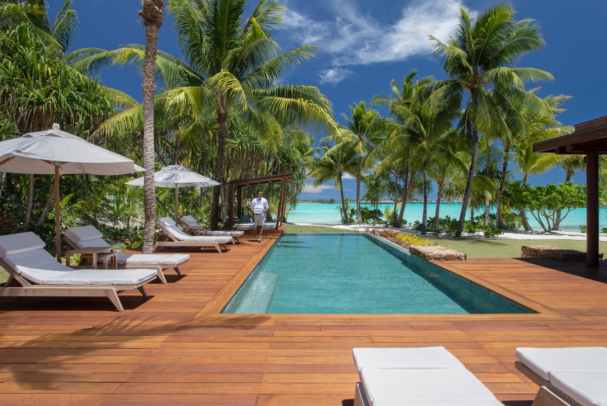 A rectangular lap pool sits centered on a warm teak wood deck lined with white cushioned lounge chairs and umbrellas, with a single person walking along the far edge. Tall coconut palms frame the space, and a white sand beach and vivid turquoise lagoon are visible through the trees in the background. A shaded pergola structure anchors the right side of the deck, and the overall setting suggests a private villa or resort property in French Polynesia.
