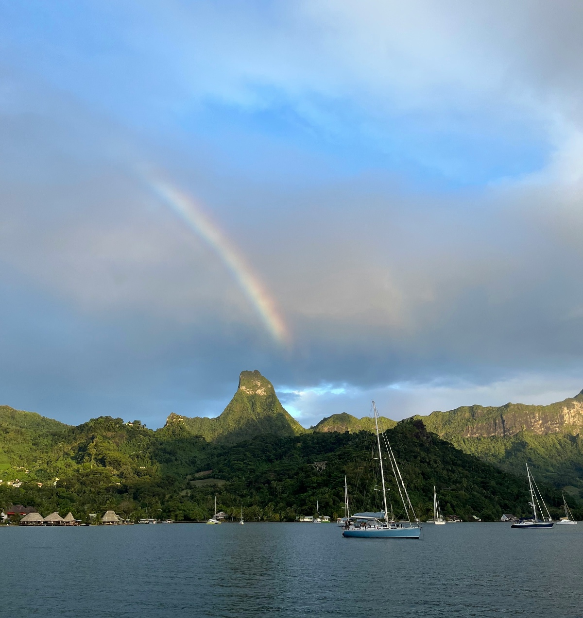 Several sailboats are anchored in a calm bay, with a dramatic volcanic peak rising sharply above densely forested green hills along the shoreline. A rainbow arcs diagonally from the upper left of the frame down to the mountain's summit, set against a partly cloudy sky with patches of blue. Thatched-roof structures are visible along the waterfront on the left, and additional boats are moored in the middle and right distance.