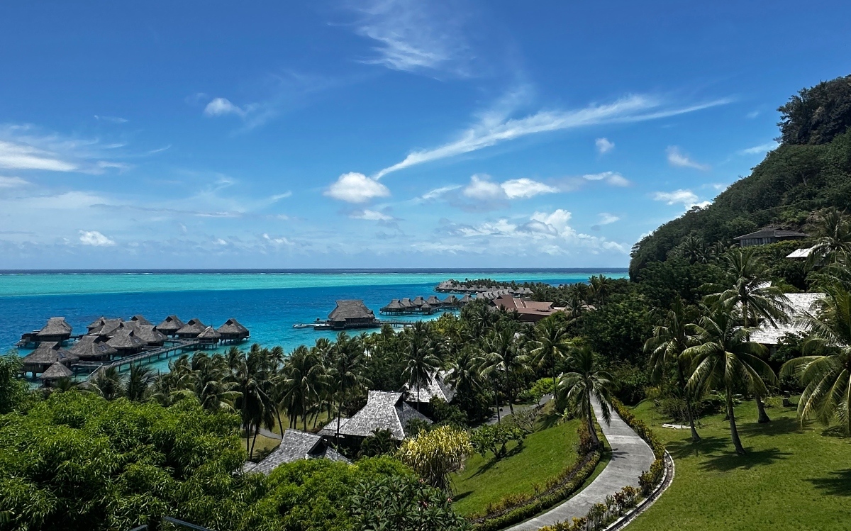 An elevated view of a French Polynesian resort shows thatched-roof bungalows nestled among dense palm trees and manicured lawns, with a curved pathway winding through the grounds in the foreground. Multiple rows of overwater bungalows extend into a vivid turquoise lagoon on the left and center, connected by wooden walkways. A densely forested hillside rises on the right, and the lagoon meets the deep blue open ocean under a bright sky with scattered clouds.