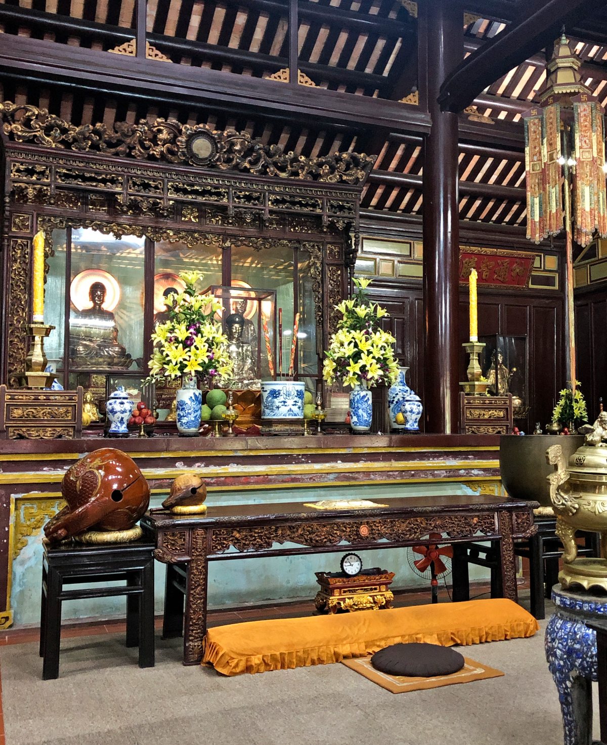 The ornate interior of a Vietnamese Buddhist temple features a gilded altar displaying Buddha statues, blue-and-white ceramic vases filled with yellow lilies, and offerings of fruit, framed by intricately carved dark wood panels. In the foreground, a carved wooden prayer table holds a wooden fish drum and mallet, with a saffron-cushioned prayer bench and meditation cushion positioned on the floor below.