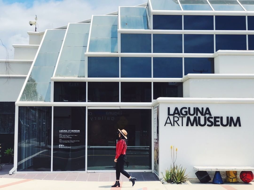 The white modern exterior of the Laguna Art Museum features a striking angular glass facade, with bold black signage on a white wall to the right. A woman in a red top, black pants, and a straw hat strides past the entrance, with small sculptural objects and desert plantings lining the base of the building.