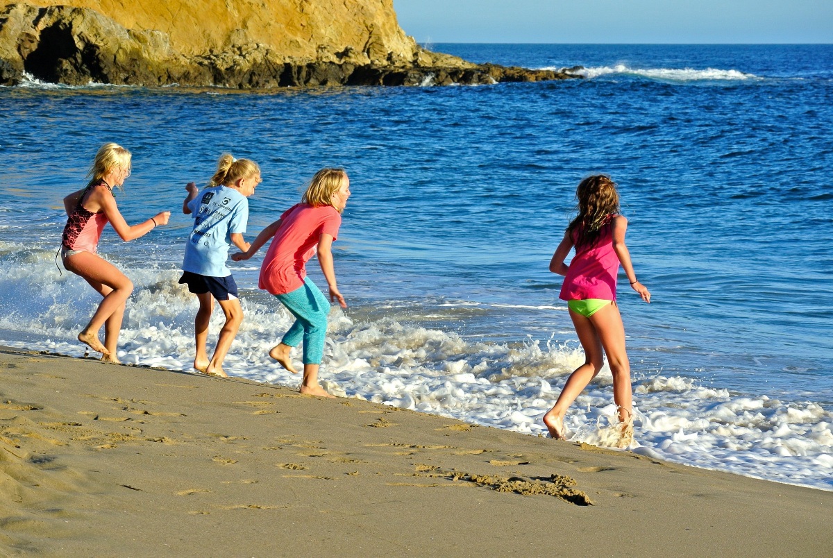 Four children run along the shoreline, stepping into shallow ocean waves on a sandy beach. A rocky cliff rises in the background under a clear sky as the water splashes around their feet.